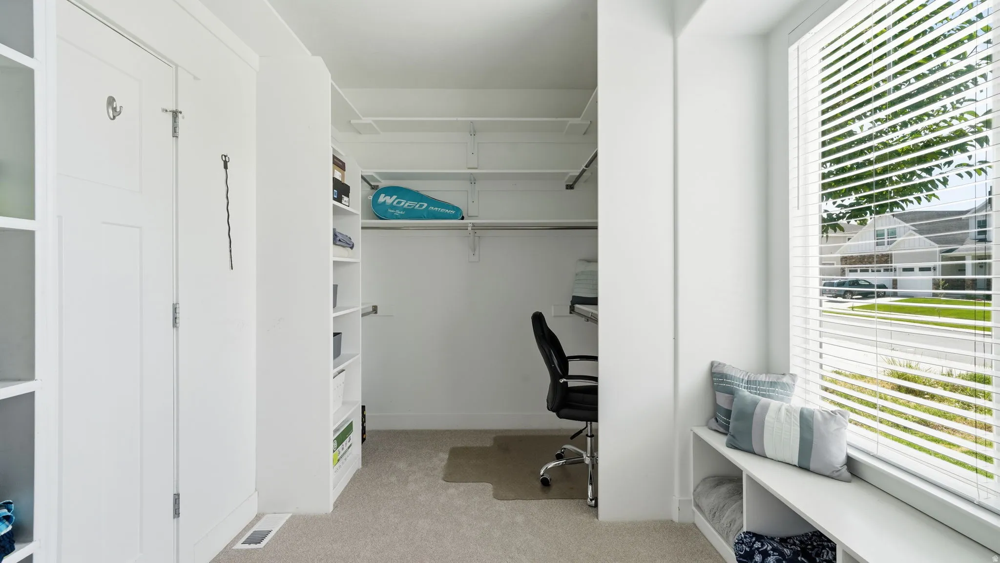 White built-in cabinetry and shelving, recessed closet space with adjustable shelving, window seating with integrated storage, large window with horizontal blinds, and carpeted flooring