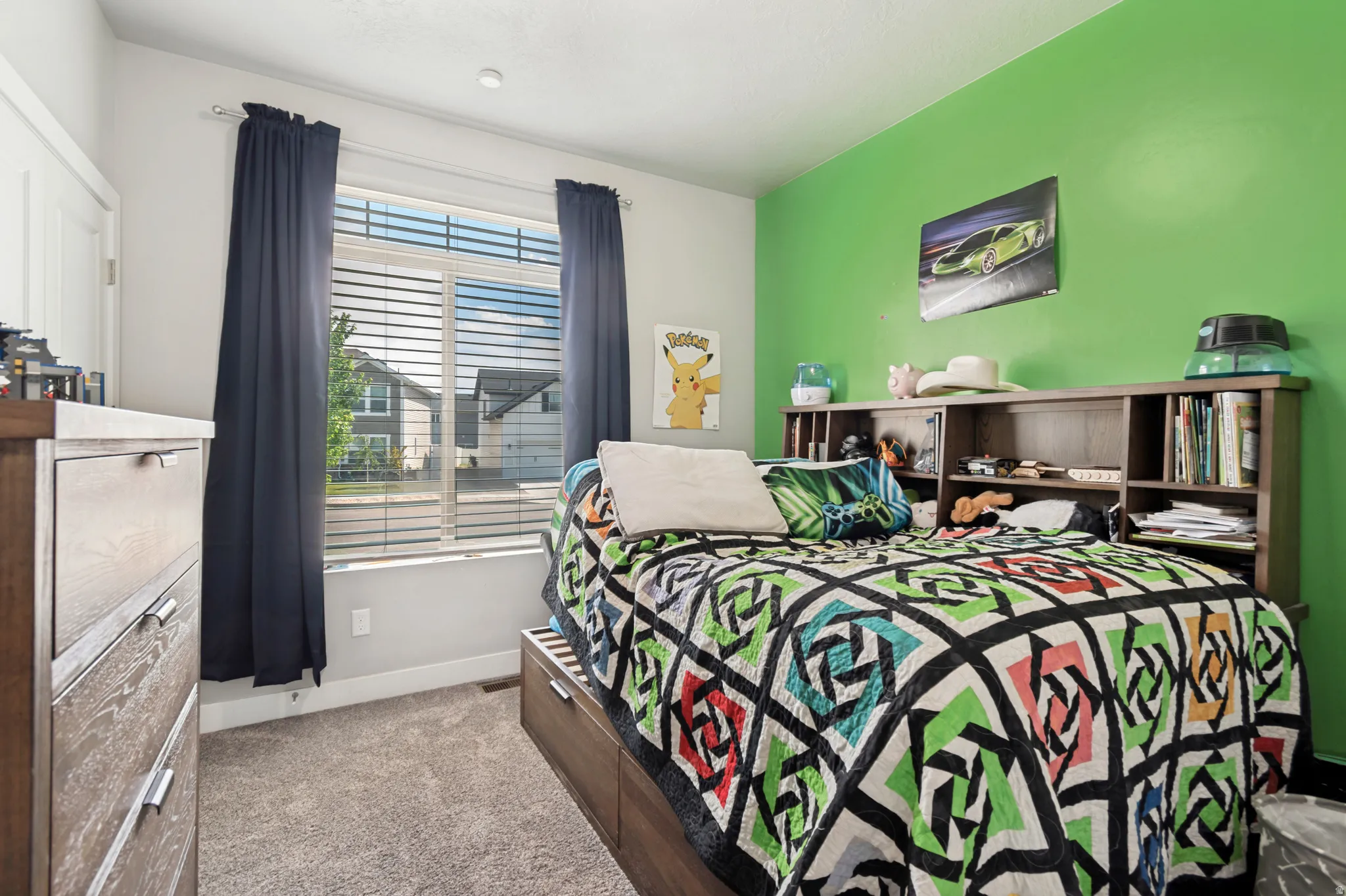 Carpeted room featuring a large window with blinds and dark curtains, a built-in bookshelf headboard, and a contrasting accent wall