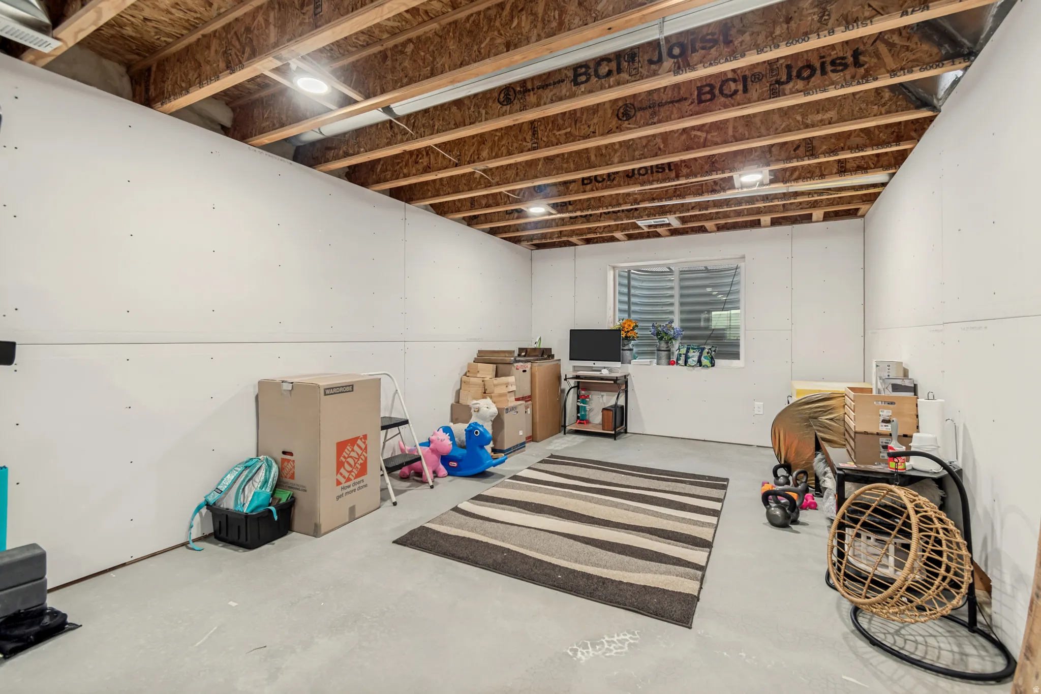 Unfinished basement space featuring exposed wood joists and subfloor, white drywall, a single egress window, and concrete flooring
