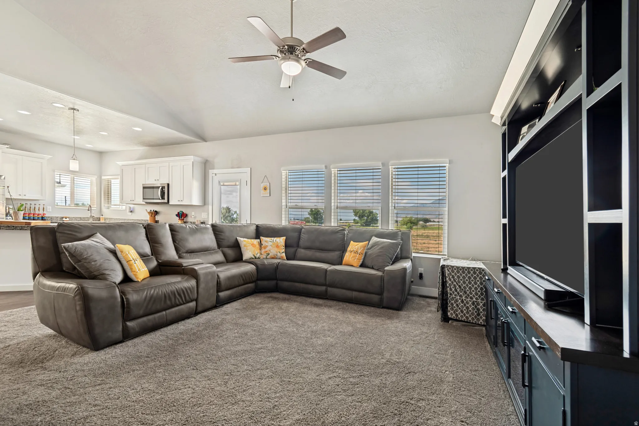Spacious living area featuring a vaulted ceiling with a ceiling fan, plush gray carpet, and a custom built-in entertainment center
