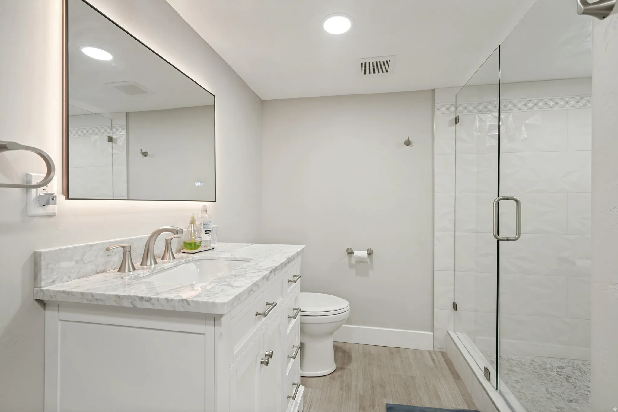 Contemporary bathroom featuring a white vanity with a stone countertop, an illuminated mirror, a glass-enclosed shower, wood-finish flooring, and recessed lighting