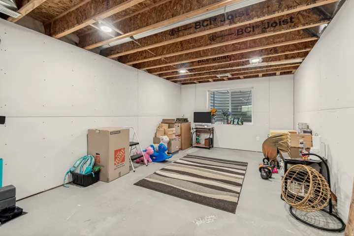 Unfinished basement space featuring exposed wood joists and subfloor, white drywall, a single egress window, and concrete flooring