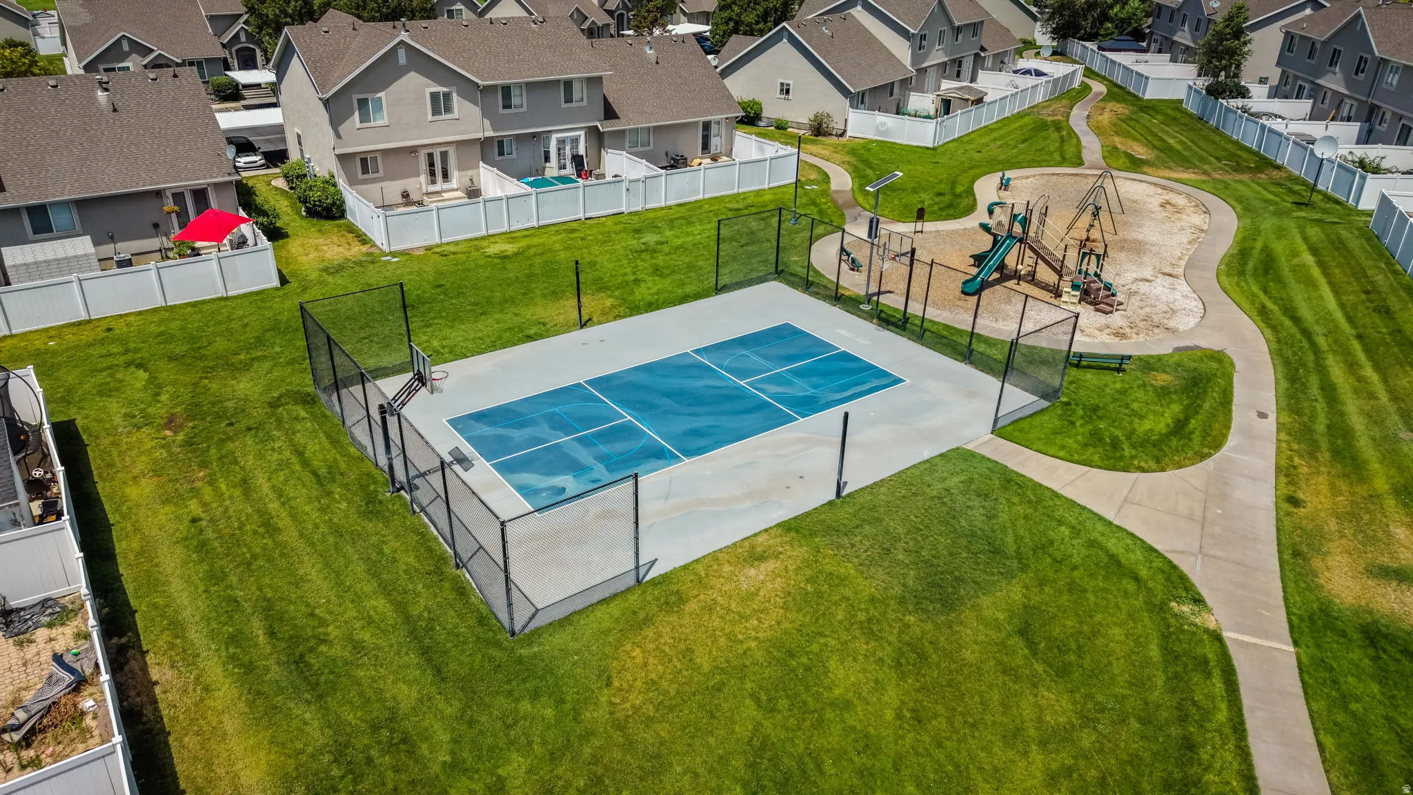 Recreational court featuring a blue playing surface, black perimeter fencing, and an adjacent playground with a slide and swing set