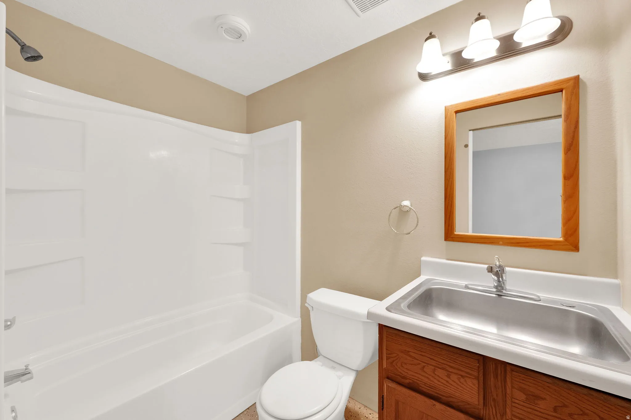 Full bathroom featuring a white tub and shower combination with integrated shelving, a wood-finish vanity with a stainless steel sink, a matching wood-frame mirror, and a three-bulb vanity light fixture