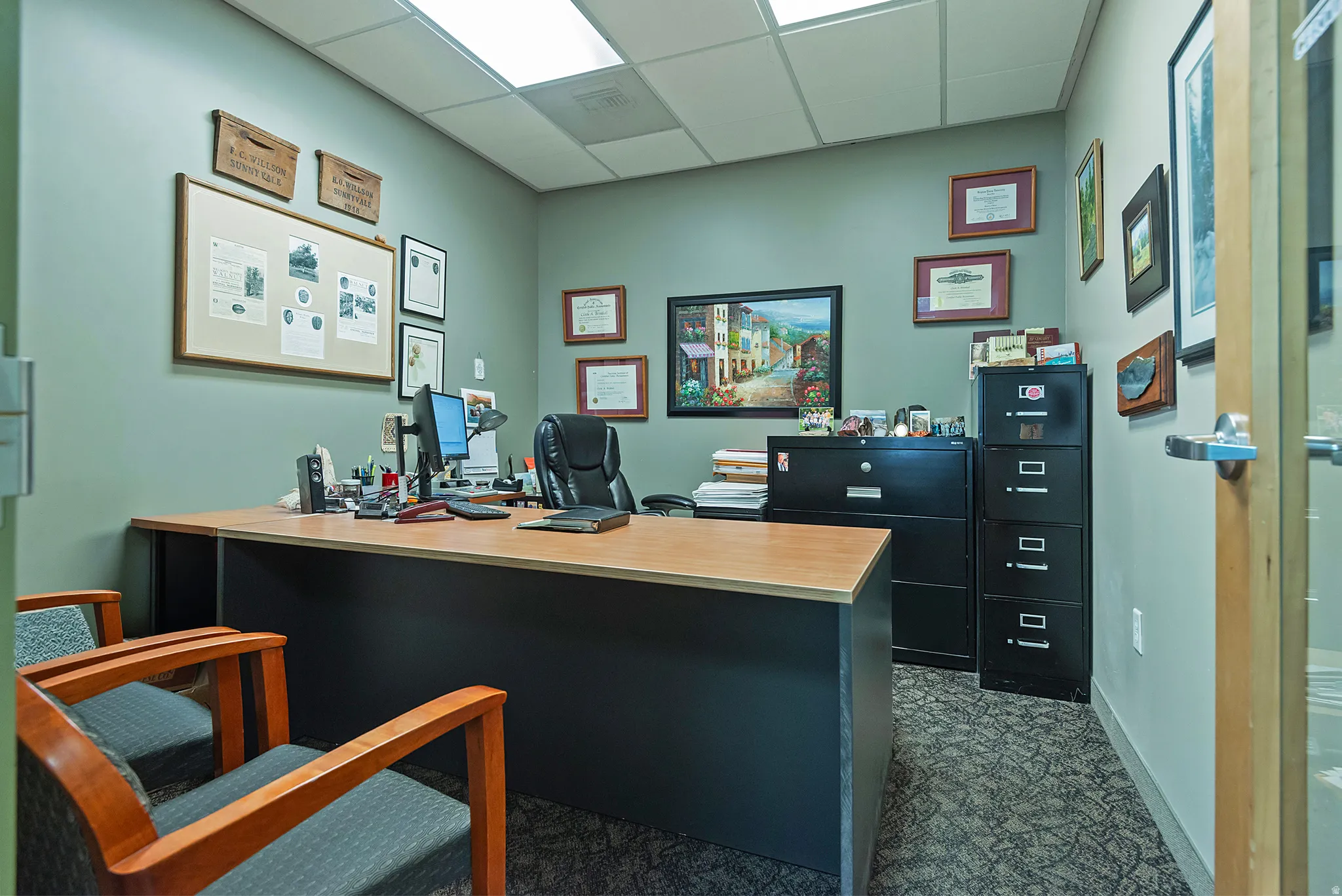 Office space featuring a suspended acoustic tile ceiling with fluorescent lighting, carpet flooring, and a built-in wood-finish desk