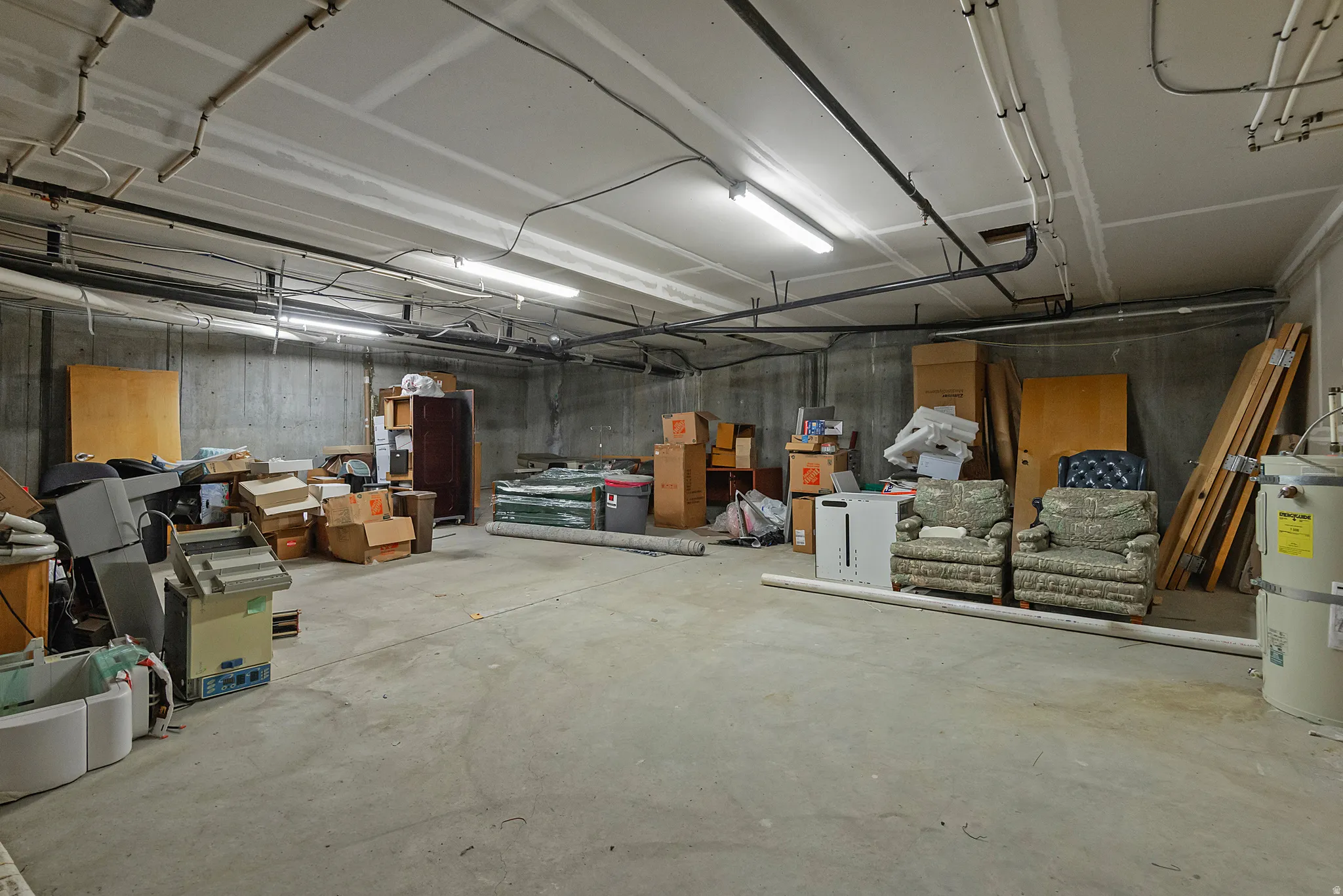 Spacious unfinished basement featuring concrete walls and flooring, exposed overhead piping, and fluorescent lighting