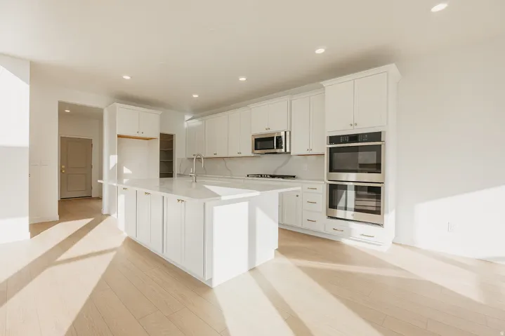 Open-concept kitchen featuring an oversized island with white cabinetry and a light-toned countertop, stainless steel appliances including double wall ovens and a microwave, recessed lighting, and light wood-finish flooring