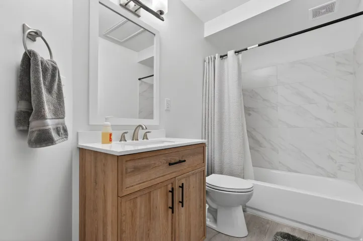 Bathroom featuring a light wood-finish vanity with a white countertop and rectangular sink, brushed nickel faucet, large framed mirror, and a white bathtub with white tiled surround