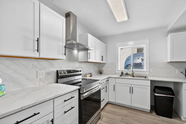Modern kitchen featuring white shaker cabinetry with black hardware, light-toned countertops, a stainless steel range with matching hood vent, and wood-finish flooring