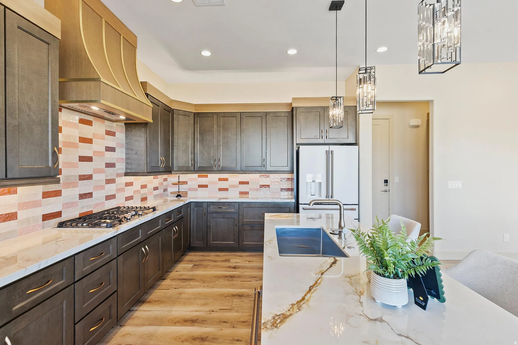 Spacious kitchen featuring an extended island with a built-in sink, stone countertops, wood-finish flooring, a decorative tile backsplash, and dark cabinetry with brass-finish hardware