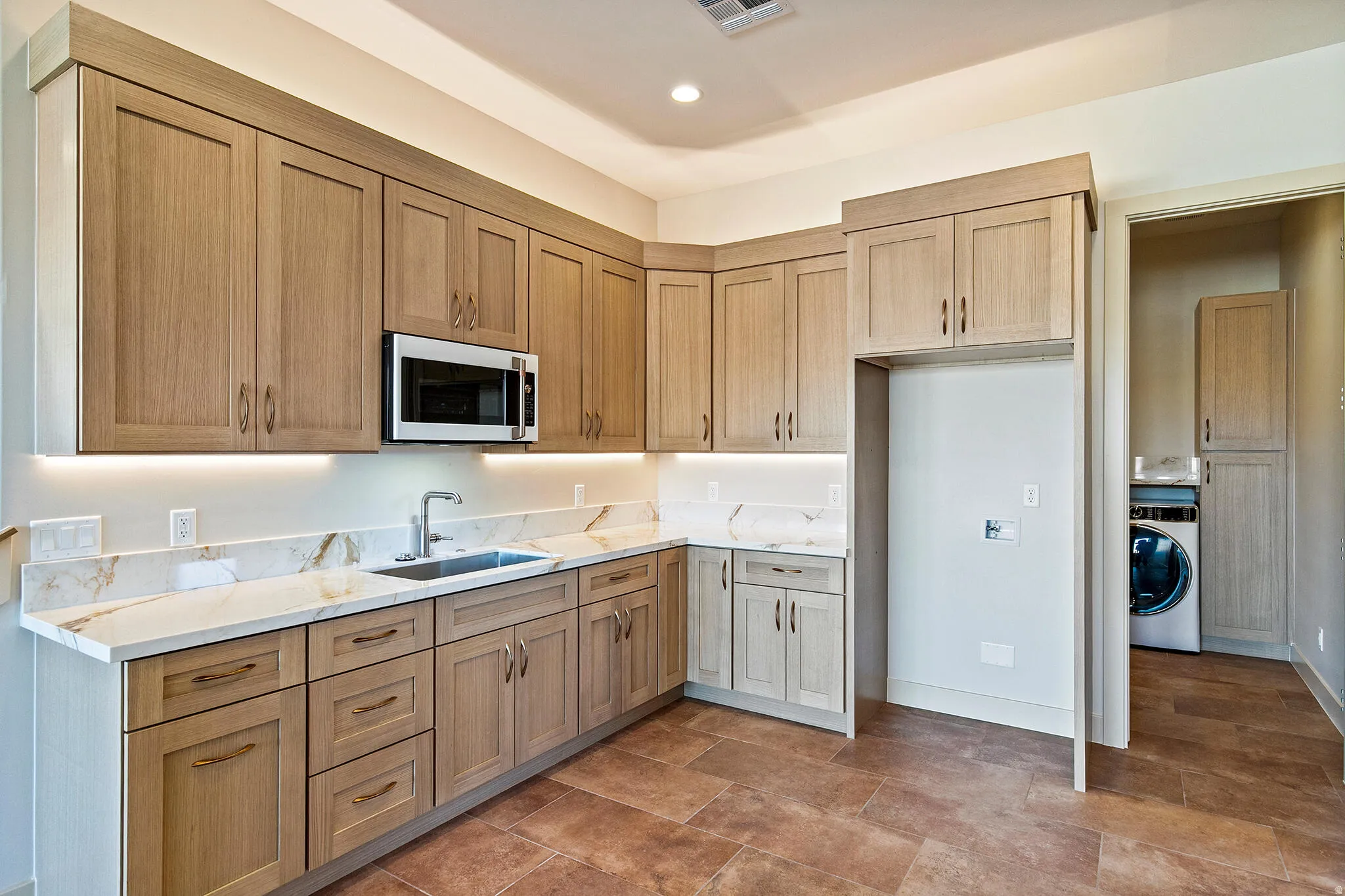 Kitchenette featuring light wood-finish cabinetry, under-cabinet lighting, a stainless-steel microwave, and light-toned countertops with an integrated sink