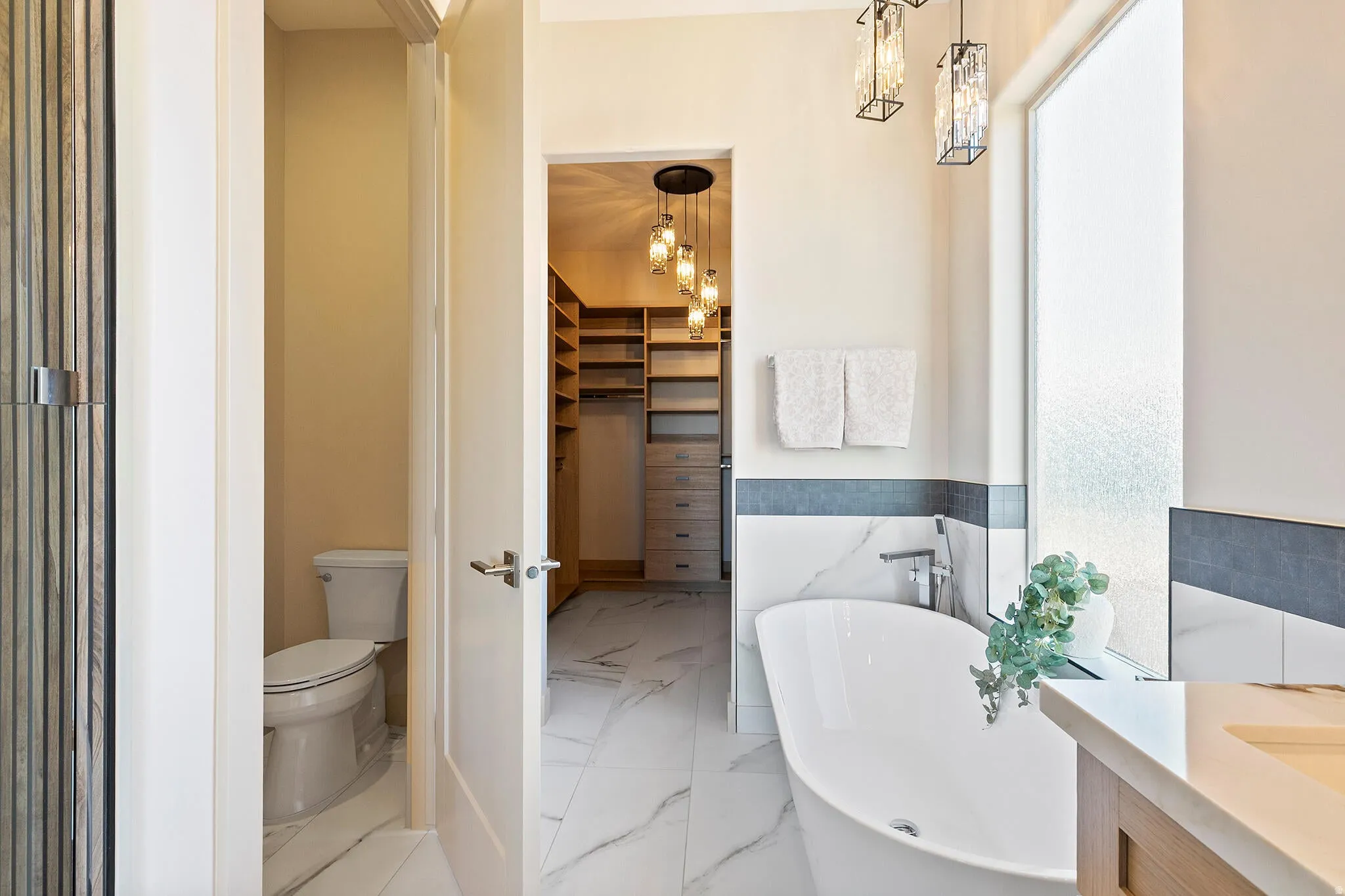 Freestanding soaking tub with chrome fixtures, framed by marble-look wall tiling and a frosted privacy window