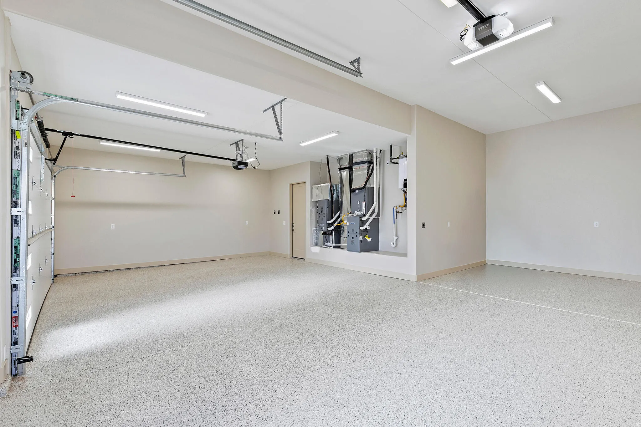Spacious garage featuring a durable epoxy floor coating, multiple overhead lighting fixtures, exposed garage door mechanisms, and a utility area with visible mechanical systems