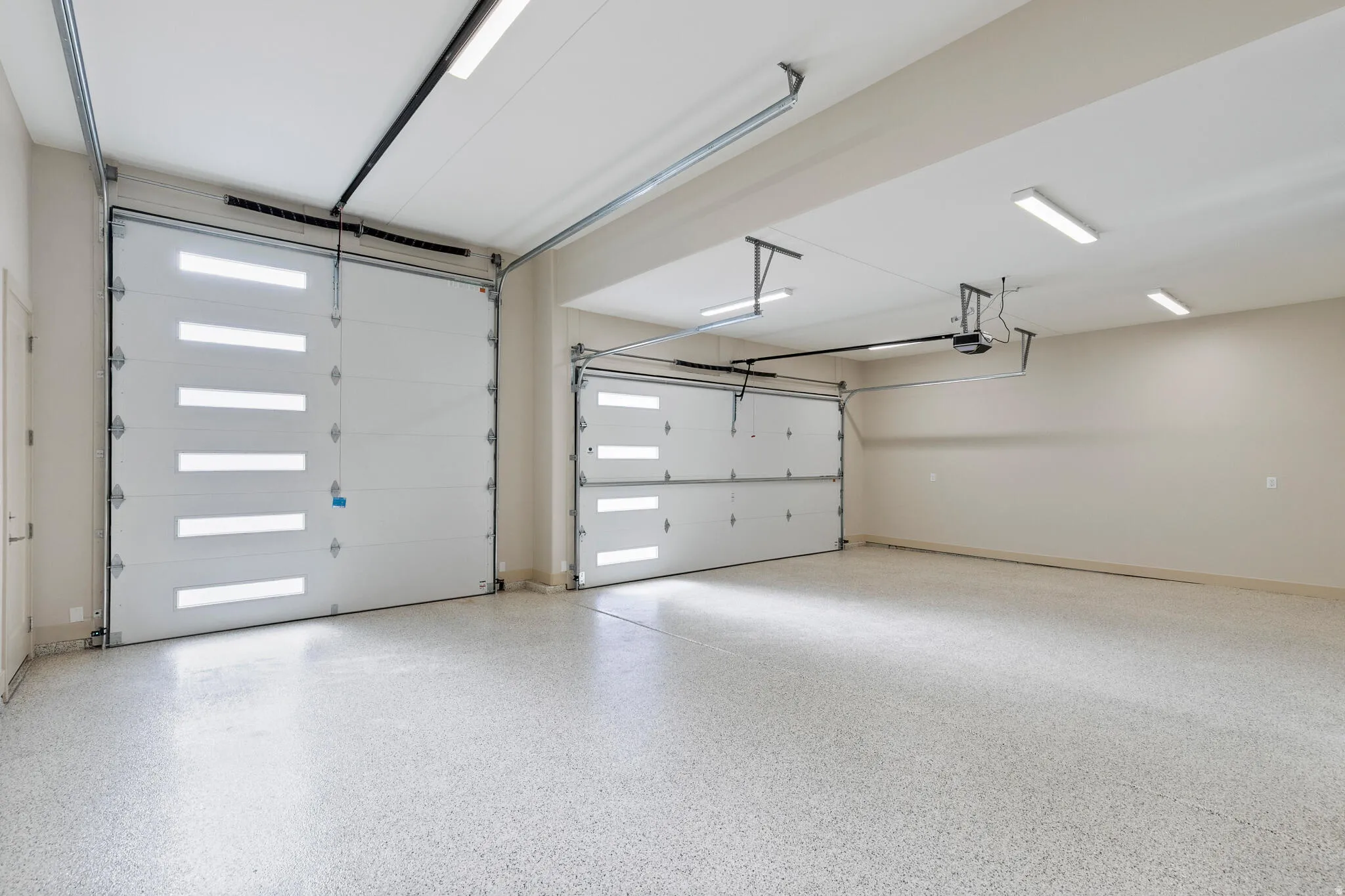 Spacious garage featuring two overhead doors with integrated light panels, an epoxy-coated concrete floor, and multiple ceiling-mounted fluorescent light fixtures