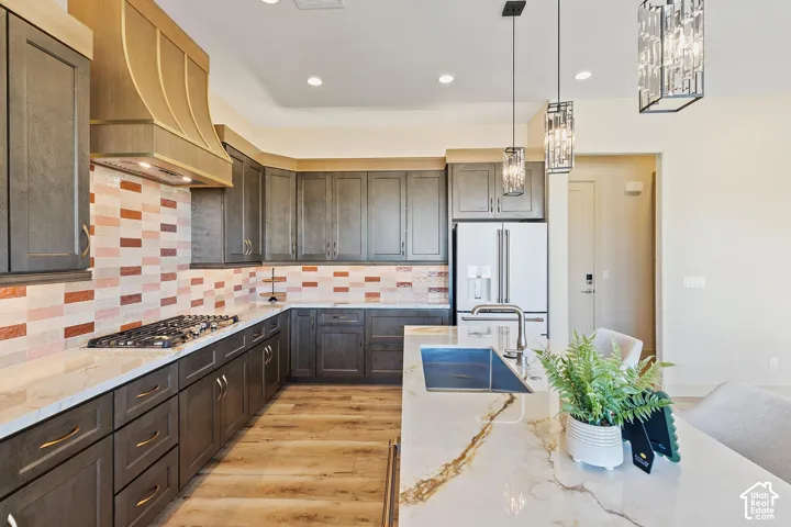 Spacious kitchen featuring an extended island with a built-in sink, stone countertops, wood-finish flooring, a decorative tile backsplash, and dark cabinetry with brass-finish hardware