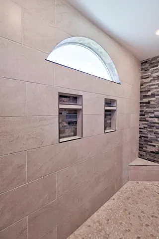 Tiled shower with a semi-circular transom window, two built-in recessed niches with accent tile, a pebble-stone floor, and a tiled bench
