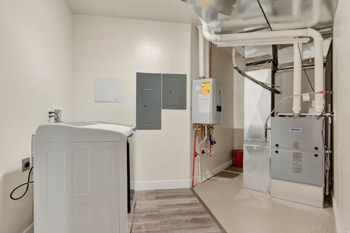 Utility area featuring a top-load washing machine, wood-finish flooring, two electrical panels, a tankless water heater, and a furnace system