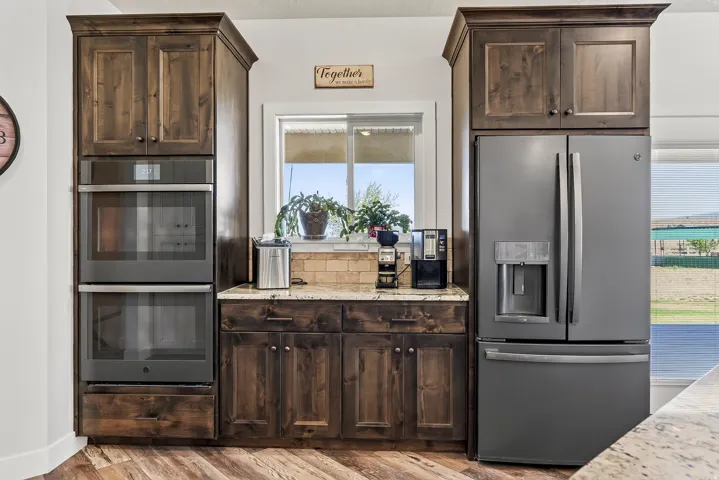 Built-in stainless steel double ovens, rich wood-finish cabinetry, light granite-style countertops, matte black refrigerator, and wood-finish flooring