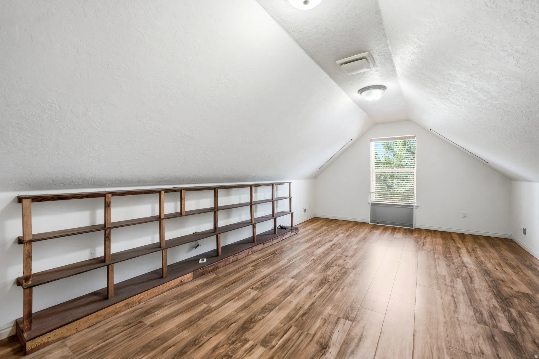 Attic space featuring wood-finish flooring, a built-in shelving unit, and a window with blinds