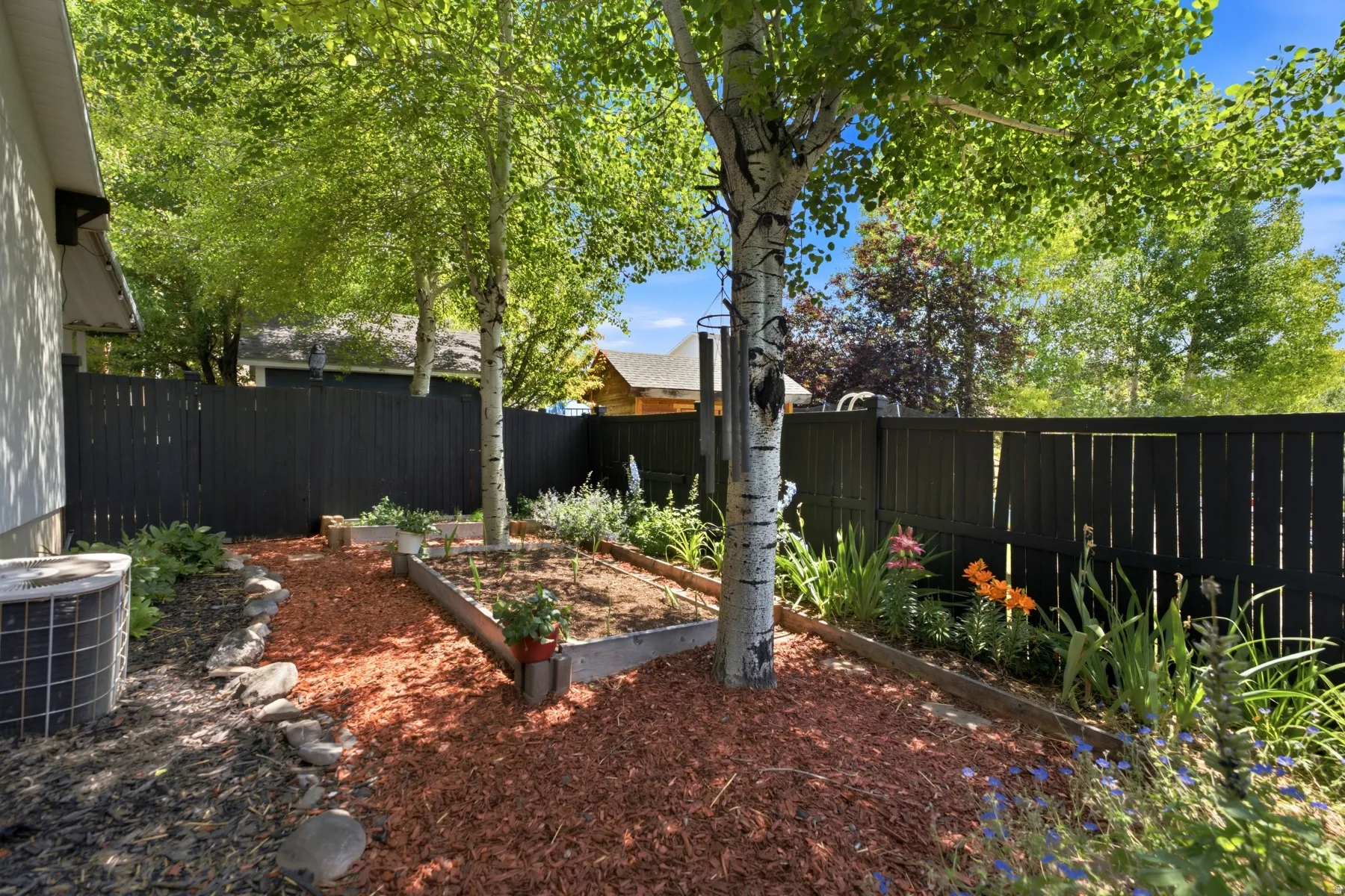 Private backyard featuring a dark-toned privacy fence, mature trees providing shade, raised garden beds, wood chip ground cover, and various planted flowers
