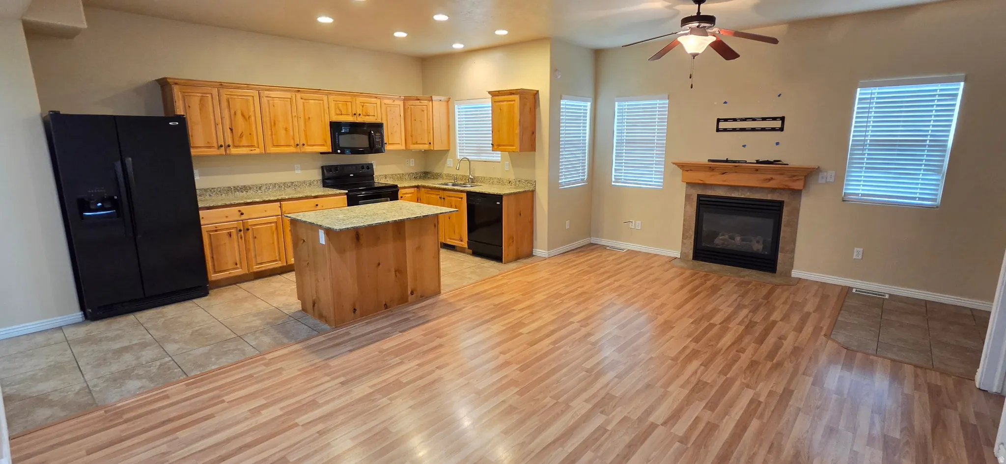 Open-concept living space featuring wood-finish flooring, a kitchen island with stone-finish countertop, black appliances, a fireplace with a wood mantel, and a ceiling fan