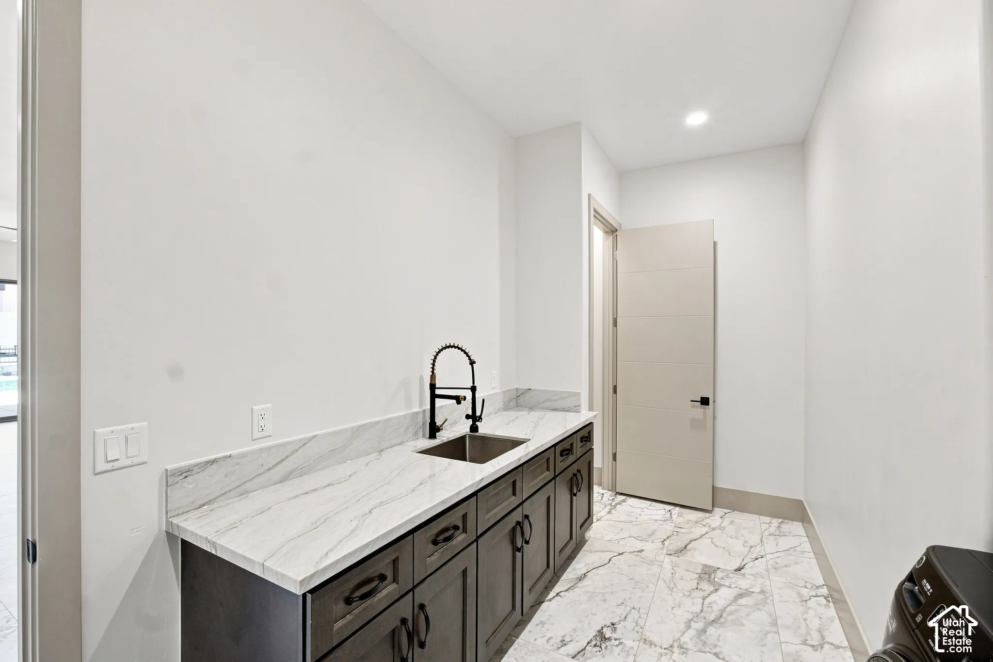 Integrated utility sink with a matte black gooseneck faucet, white-veined countertop, dark wood-finish cabinetry, and large format floor tiling