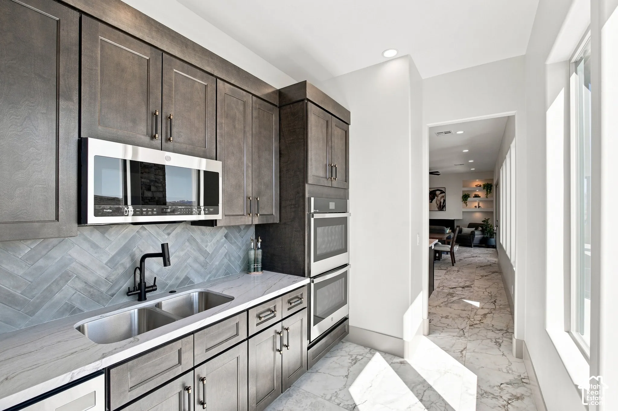 Contemporary gray cabinetry, dual stainless steel ovens, stainless steel microwave, dual basin undermount sink with a matte black faucet, and white countertop with a gray herringbone tile backsplash