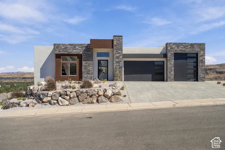 Modern architectural exterior with stone veneer accents, integrated garage doors, a concrete driveway, and natural rock landscaping