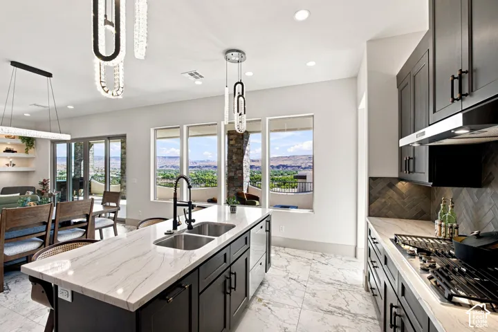 Kitchen island with dual basin sink and stone countertop, dark cabinetry with matte black hardware, herringbone tile backsplash, and large windows
