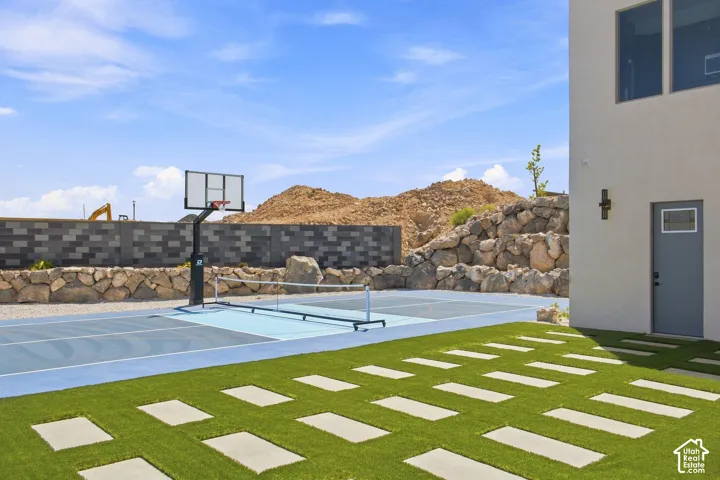 Multi-sport court with a basketball hoop and net, artificial turf patio featuring rectangular stepping stones, and a stucco exterior with a gray door