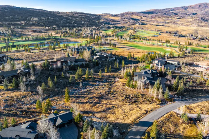 Expansive mountain landscape featuring multiple custom homes, a winding paved road, scattered evergreen trees, and distant green golf course fairways with a water feature