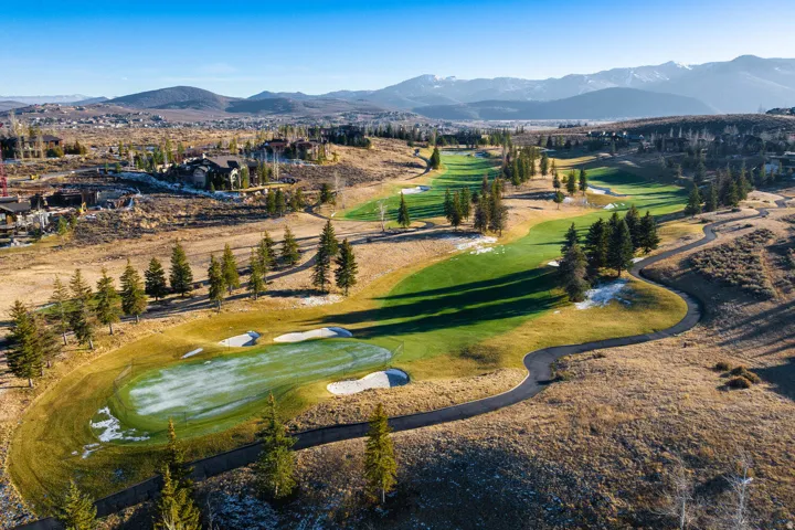 Expansive golf course terrain featuring manicured greens, sand traps, paved cart paths, scattered evergreen trees, and distant mountain ranges