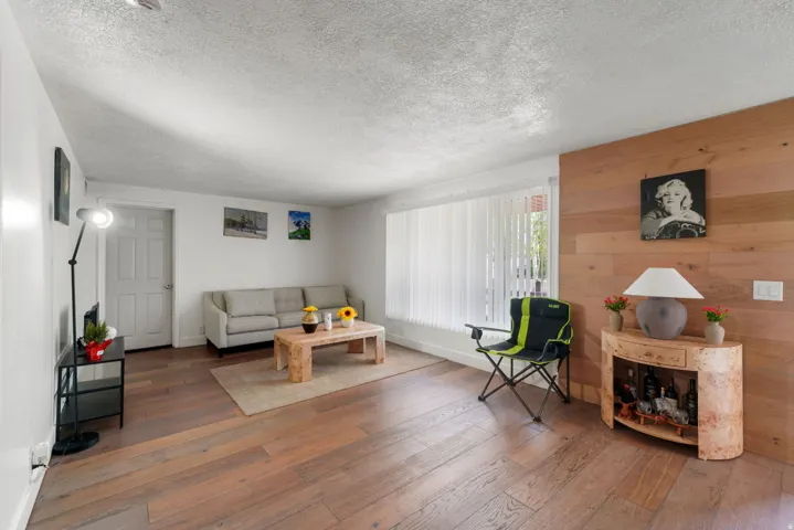 Spacious living area featuring wide plank wood-finish flooring, a prominent wood-paneled accent wall, and a large window with vertical blinds