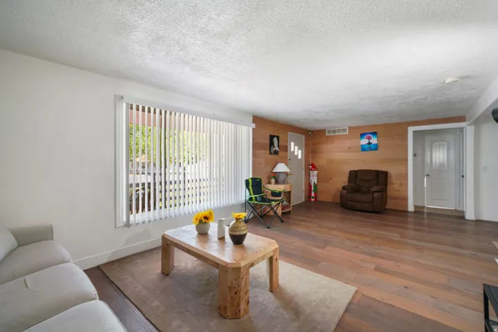 Living area featuring wood-finish flooring, a full wall with horizontal wood paneling, and a large window with vertical blinds