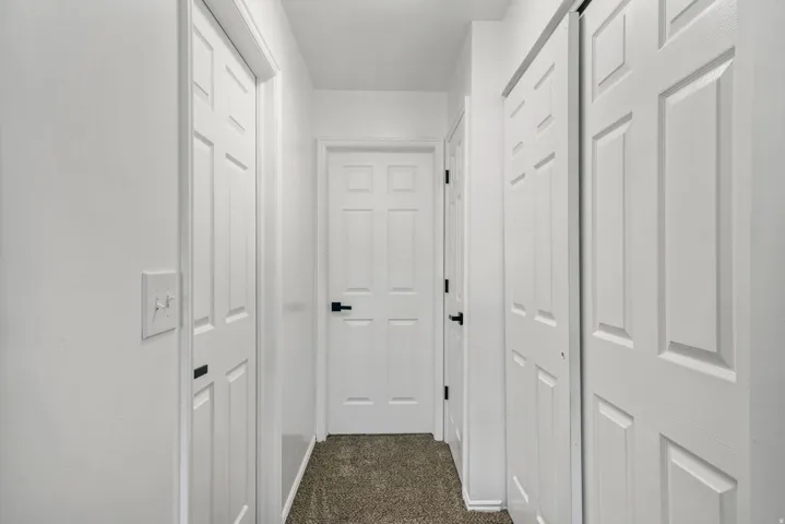 Hallway featuring white paneled doors, neutral wall paint, overhead lighting, and dark textured carpeting