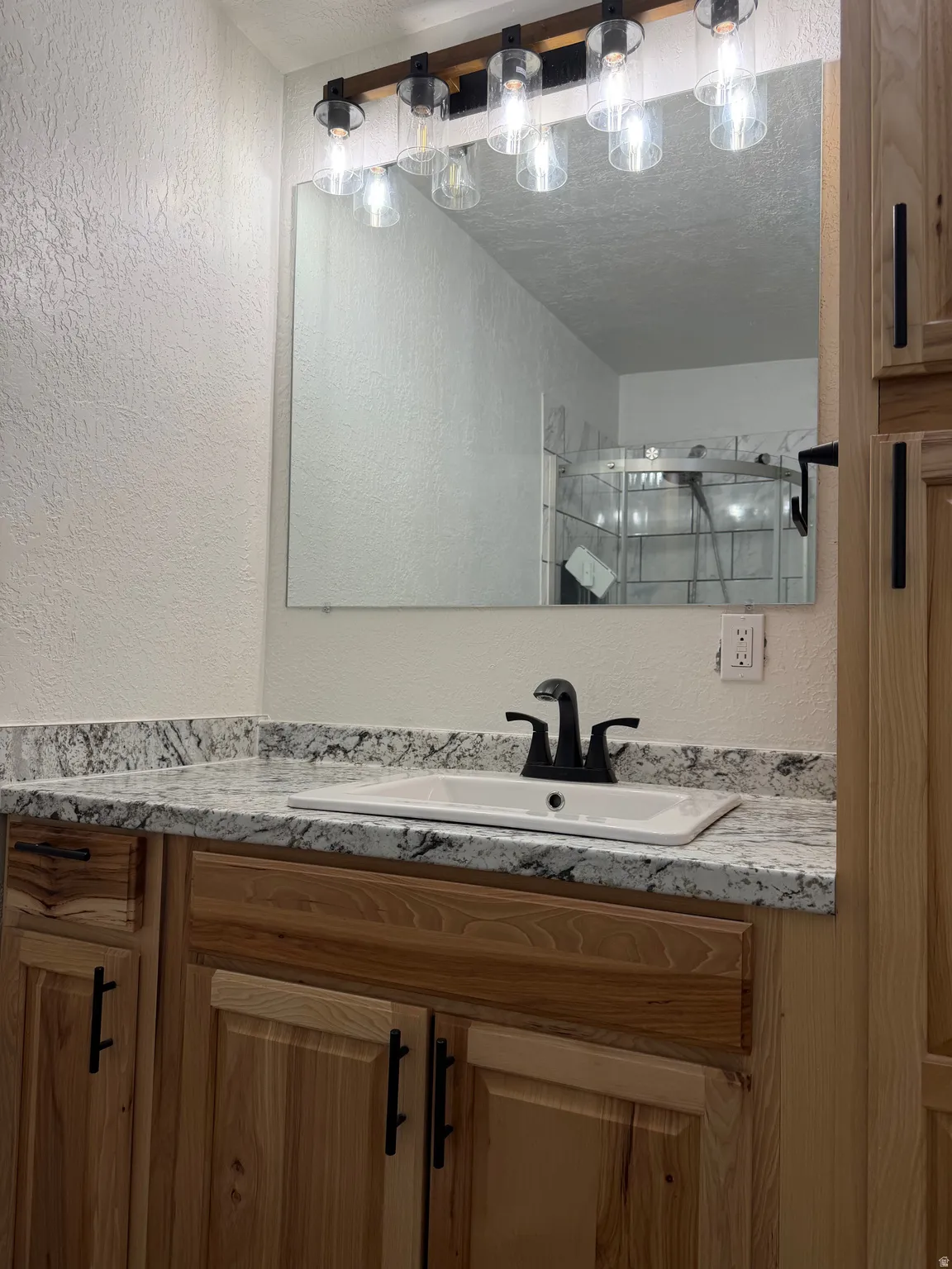Bathroom vanity featuring natural wood cabinetry, a speckled countertop, an undermount sink with a matte black faucet, and a contemporary five-light fixture