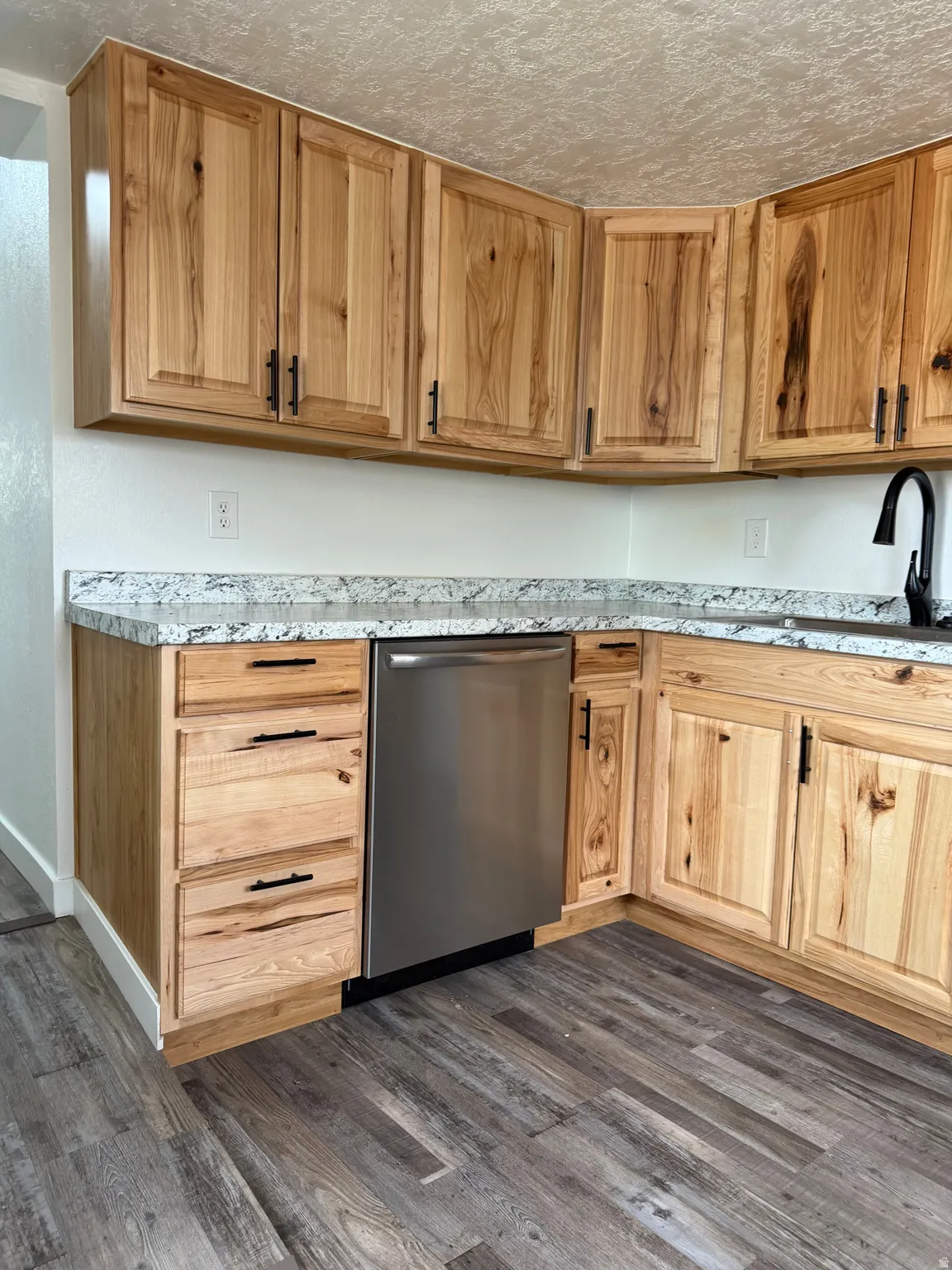 Kitchen featuring natural wood cabinetry with black hardware, light-toned countertops, a stainless steel dishwasher, and wood-finish flooring
