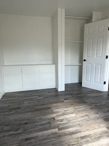 Room featuring wood-finish flooring, built-in storage drawers, an open closet with shelving, and a paneled interior door with black hardware