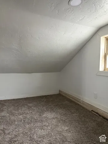 Attic space featuring sloped ceilings, textured white walls, a single-pane window with wood trim, and neutral-toned carpeting
