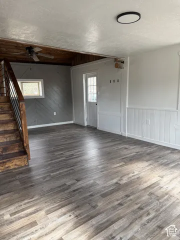 Spacious interior featuring wood-finish flooring, a built-in wooden staircase with dark metal railing, a ceiling fan, and white paneled wainscoting