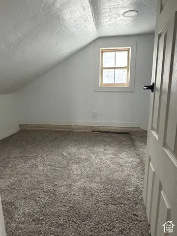 Carpeted attic space featuring sloped ceilings, a single-pane window with wood trim, a flush-mount ceiling light, and a paneled door with a black handle