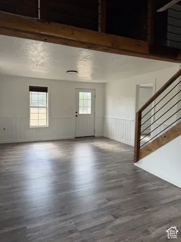 Spacious room featuring wood-finish flooring, white paneled wainscoting, an exposed wood beam ceiling, and a contemporary staircase with black metal balusters