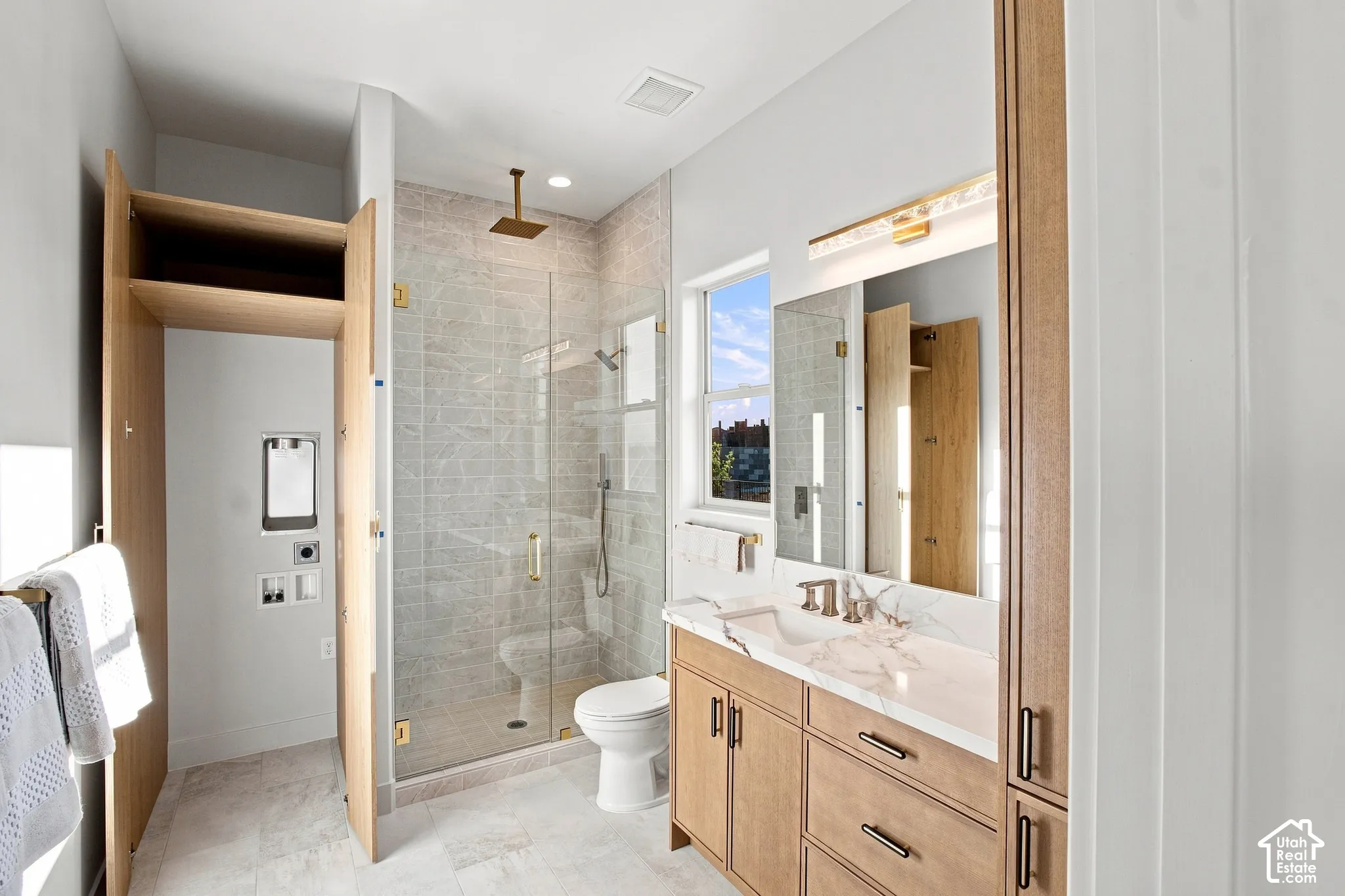 Spacious bathroom featuring a glass-enclosed shower with a rainfall showerhead, a single vanity with a stone countertop, wood-finish cabinetry, and a dedicated laundry alcove