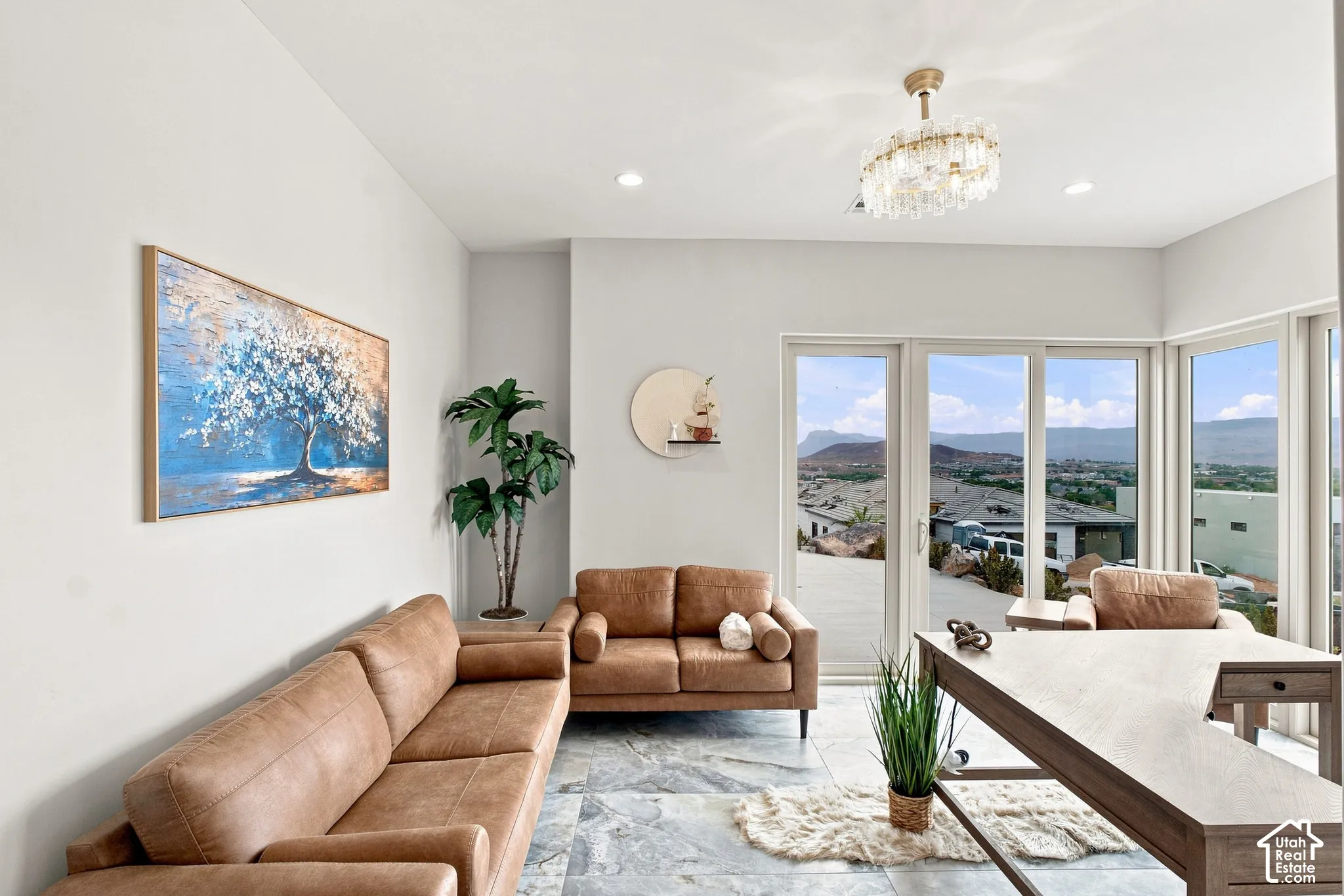 Sophisticated living space showcasing floor-to-ceiling multi-panel glass doors and windows framing expansive landscape vistas, light-filled interiors with recessed ceiling lighting, a crystal chandelier, neutral wall finishes, and polished stone flooring