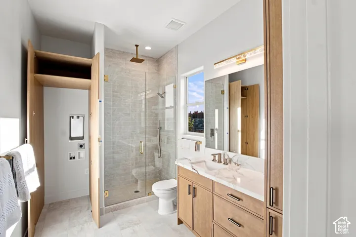 Spacious bathroom featuring a glass-enclosed shower with a rainfall showerhead, a single vanity with a stone countertop, wood-finish cabinetry, and a dedicated laundry alcove