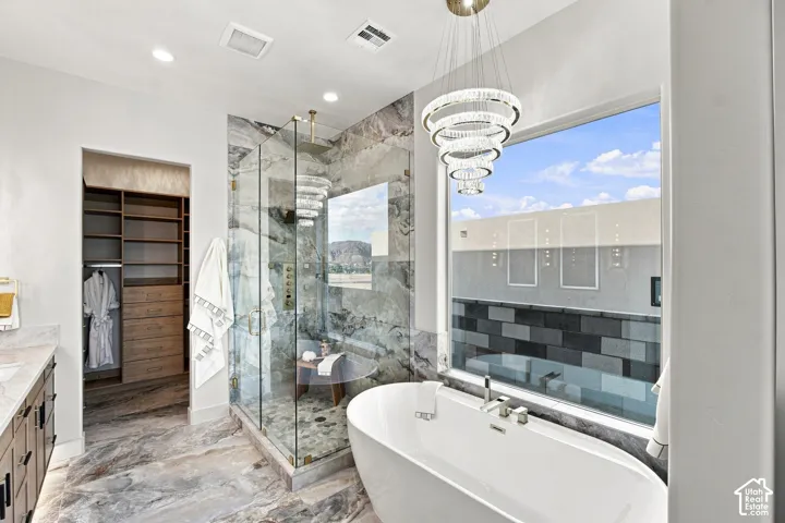 Freestanding soaking tub, glass-enclosed shower with stone-finish surround, large window, custom built-in closet system, and crystal chandelier