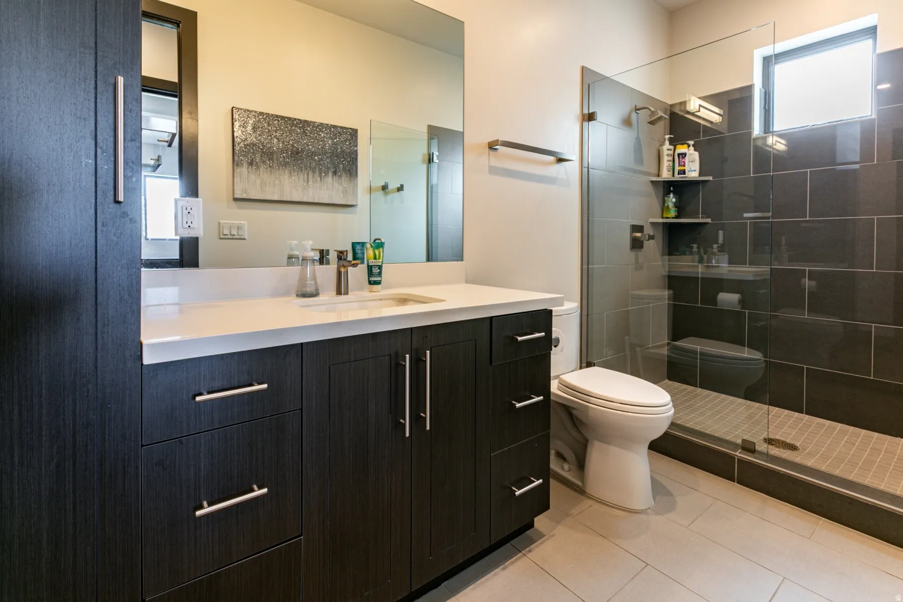 Bathroom featuring a dark wood-finish vanity with a light countertop, a glass-enclosed shower with dark rectangular wall tiles, and light-colored floor tiles