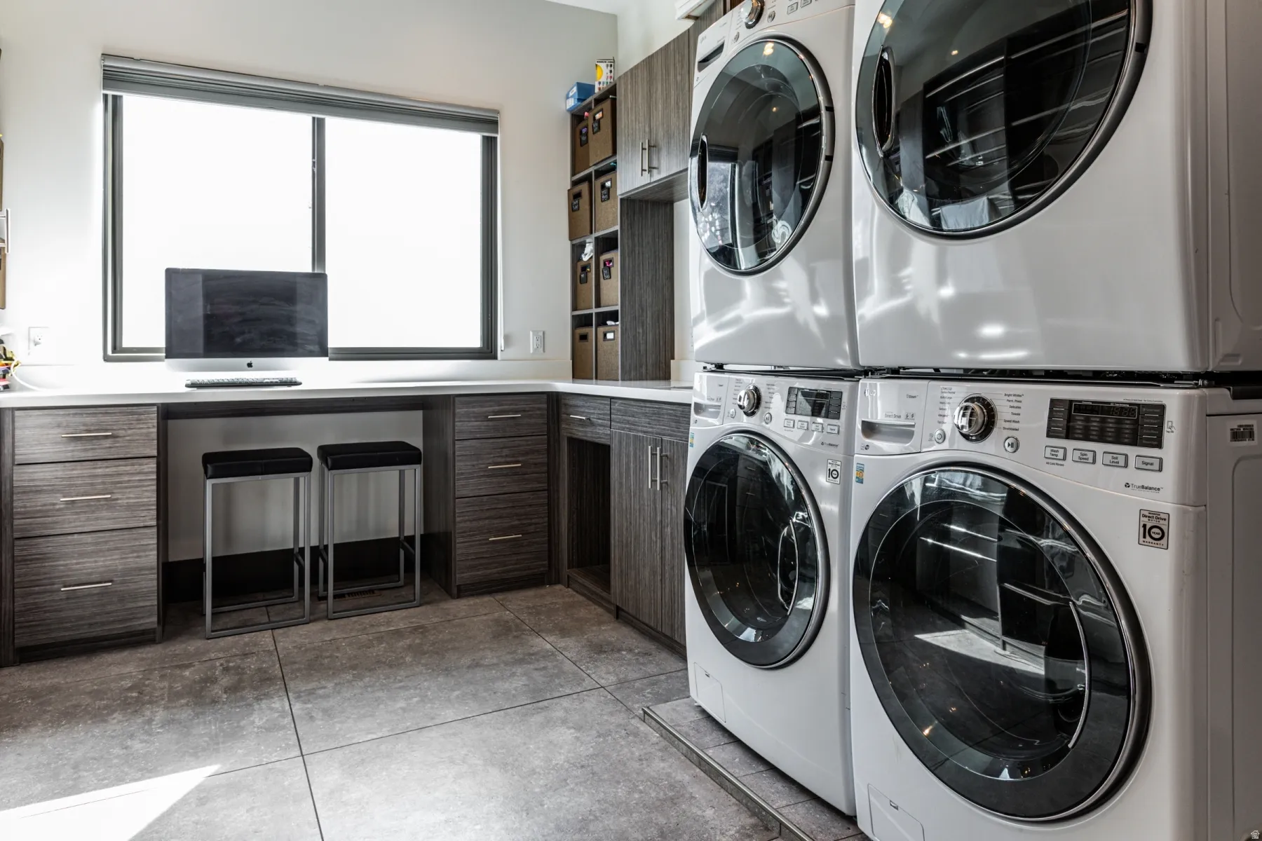 Laundry area featuring stacked front-loading appliances, a built-in desk with quartz-style countertop, textured wood-finish cabinetry, and large format tile flooring
