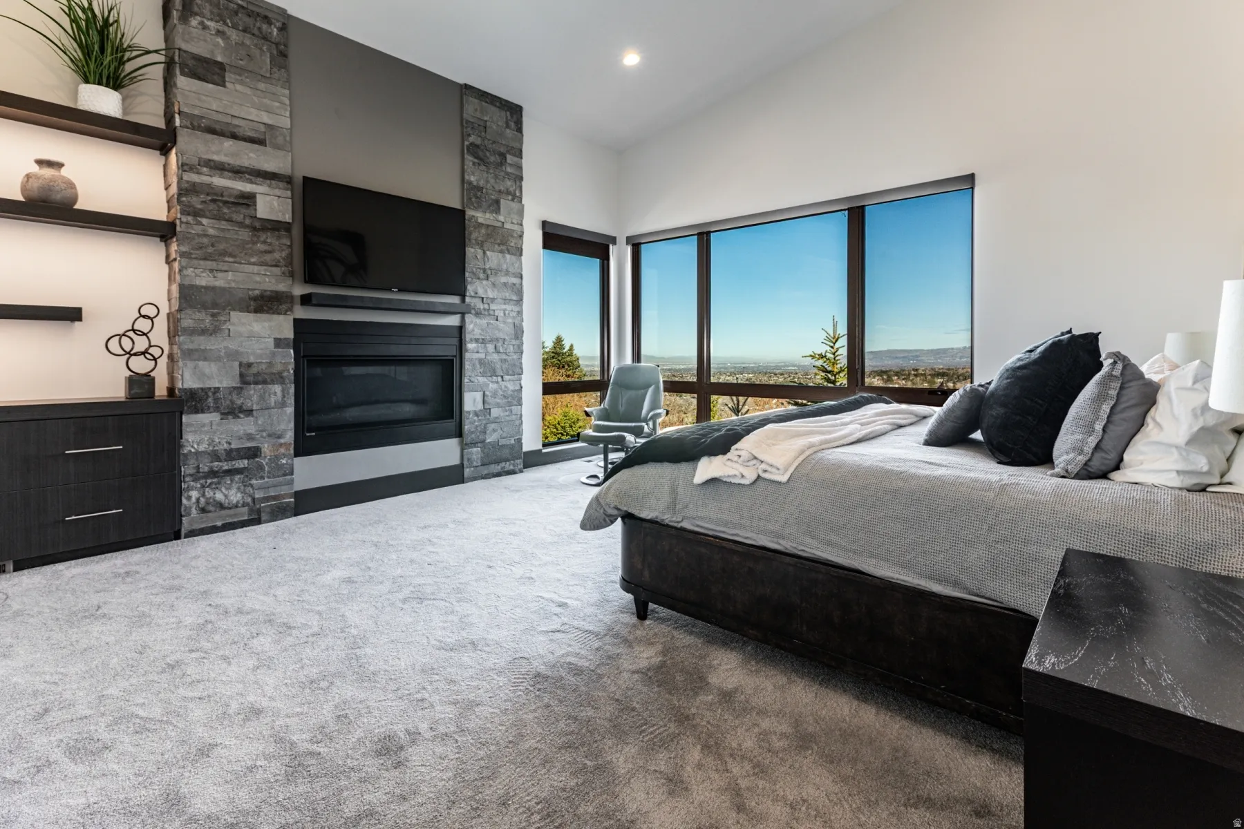 Spacious master bedroom featuring broad windows with distant scenery, a built-in fireplace with a stone surround, and light-colored carpeting