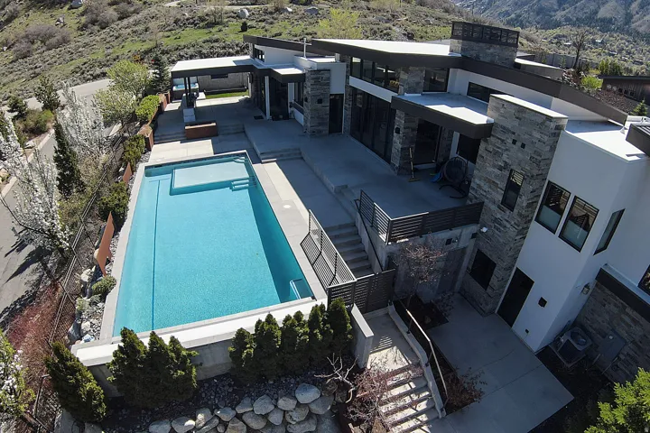Modern architectural residence featuring multi-level stone and stucco exterior, an integrated swimming pool with an elevated spa, extensive concrete patio areas, and multiple outdoor balconies